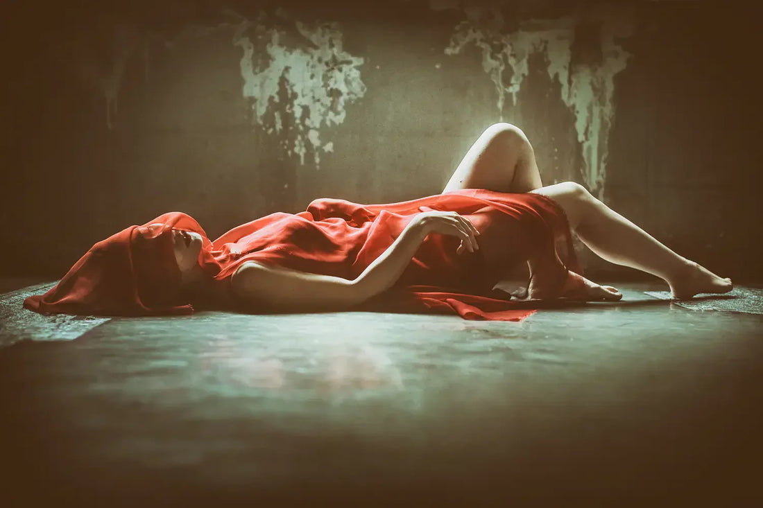 Woman lying on floor in sheer red dress with fabric covering her face in moody dimly lit room