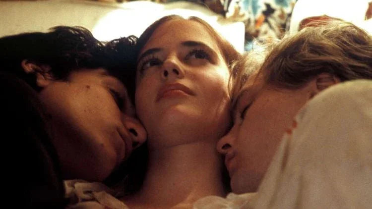 A woman lying between two men, all close together in an intimate setting, with neutral expressions.