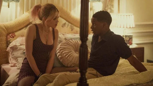 Young woman with pink hair and black dress sitting on bed talking to young man in dark shirt in cozy bedroom